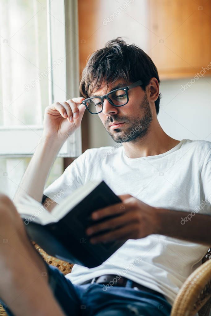Young handsome man in eyeglasses reading book sitting on chair at home