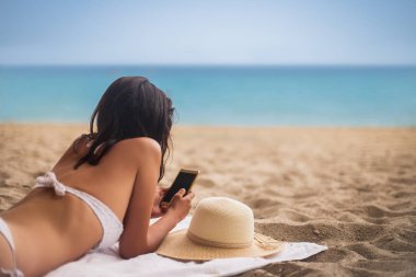 Tropik sahilde yalan ve cep telefonuyla bikinili genç güzel kadın. Yaz tatil kavramı