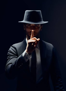 Siyah takım elbise li ve şapkalı ciddi bir adam dudaklarda parmağını tutar, sessiz jest yapar ve karanlık arka planda komployu izole eder. Shh, sessizlik kavramı                           