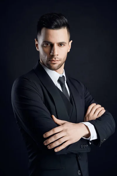 Handsome confident man in black suit with arms crossed on dark background. Young and success concept
