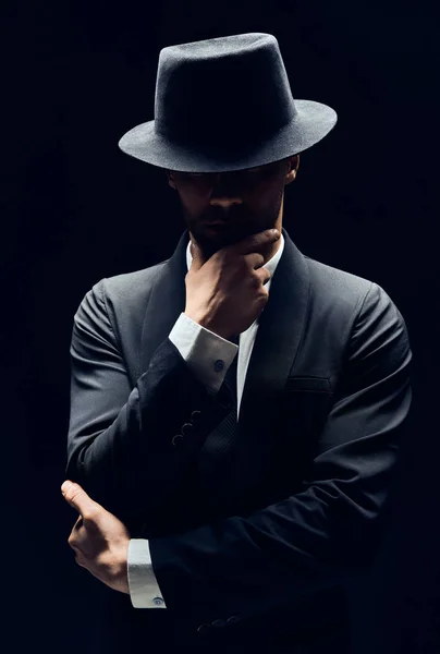 Handsome thoughtful man in black suit and hat touching chin isolated on dark background