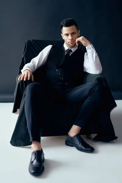 Confident thoughtful man in elegant suit posing sitting in chair on black background