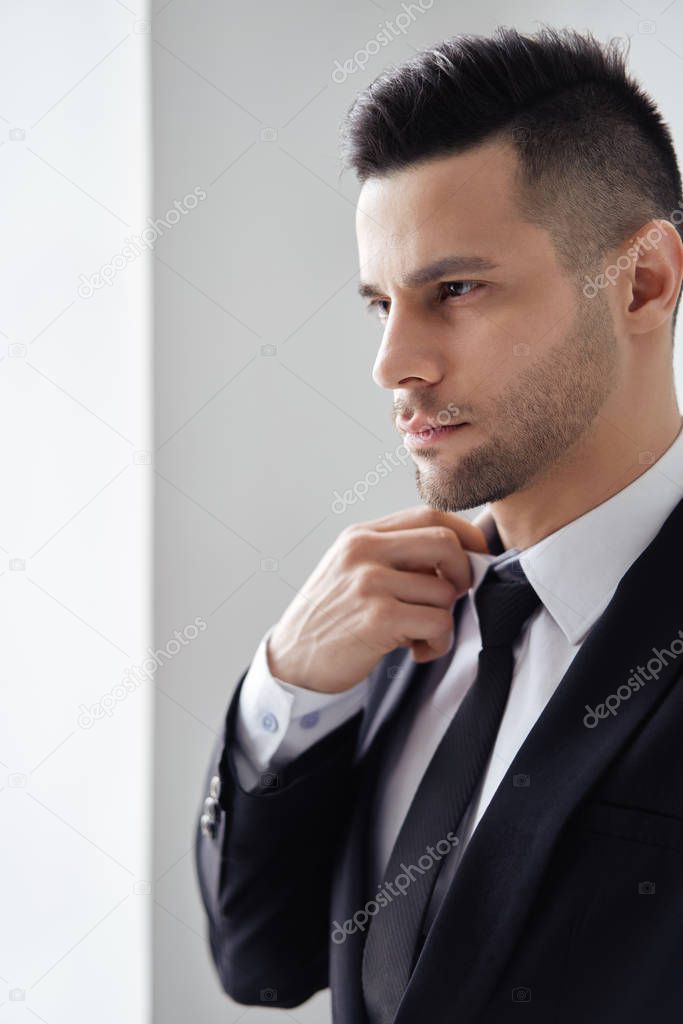 Young handsome man in elegant suit adjust his necktie on white background                       