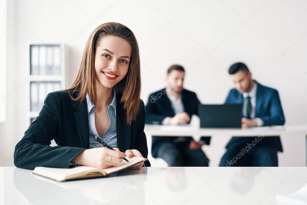 Happy smiling business woman portrait sitting in office and making notes with her business team on background. Leadership and success concept                           