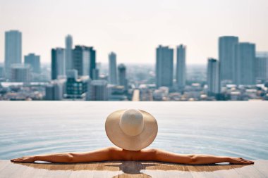 Çatıdaki yüzme havuzunda rahatlatıcı şapkalı güzel kadın, Cityscape Panorama manzarasının keyfini çıkarabilir. Tatil, yaz ve Relax konsepti