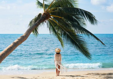 Şapka rahatlatıcı ve tropikal plajda palmiye ağacı altında deniz ayakta keyfini güzel genç kadın geri görünümü. Yaz tatili konsepti                      