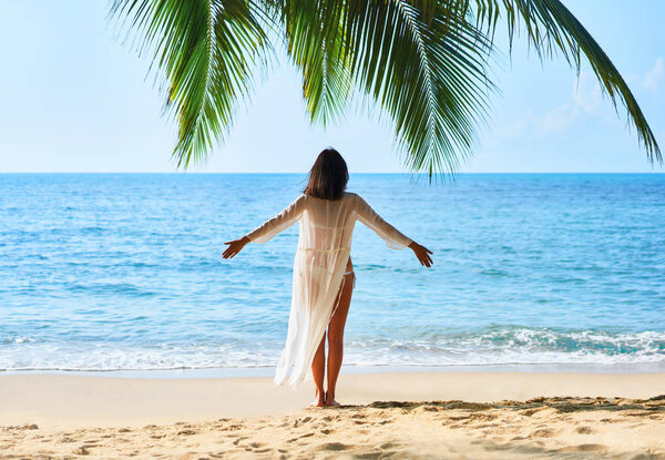 Back view of free young woman enjoying sea standing under palm tree on tropical beach.