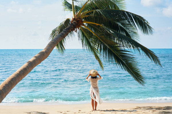 Back view of pretty young woman in hat relaxing and enjoy sea standing under palm tree on tropical beach. Summer vacation concept                      
