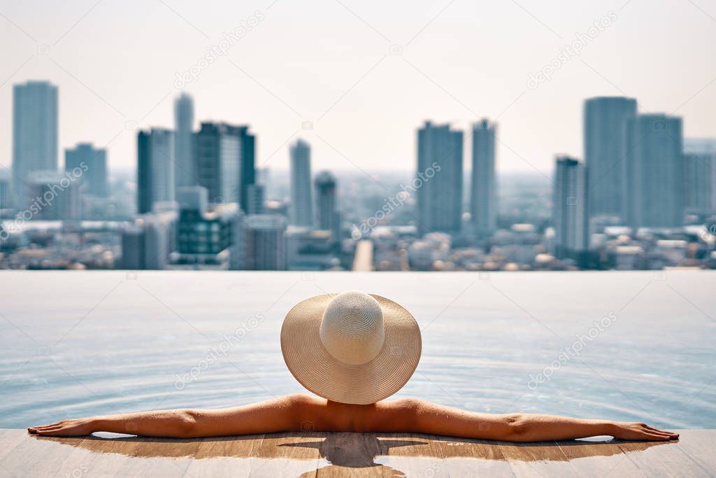 Pretty woman in hat relaxing in swimming pool on roof top enjoy cityscape panorama view. Vacation, summer and relax concept