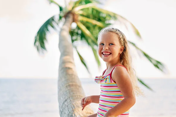 ヤシの木に座ってビーチで遊ぶ幸せな小さなブロンドの女の子 — ストック写真