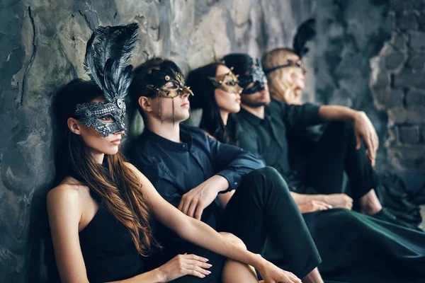 Group of friends in masquerade carnival masks sitting on floor relaxing after party. Women and men wearing venetian masks. Fashion, friends concept.