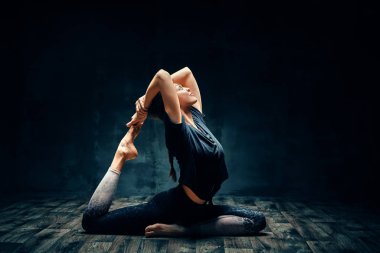 Yoga yapan genç bir kadın karanlık odada tek bacaklı kral güvercin pozu veriyor. Eka Pada Rajakapotasana asana.