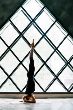 Yoga yapan genç bir kadın destekli amuda kalkıp duruyor, Salamba Sirsasana, pencere arkasında.. 