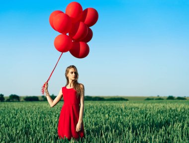 Kırmızı elbiseli genç bir kadın yeşil alanda kırmızı balonlarla dikiliyor.