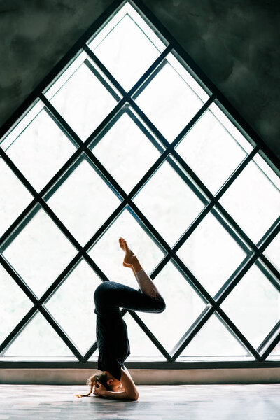 Young woman practicing yoga standing in supported headstand pose, akunchanasana, on window background. 