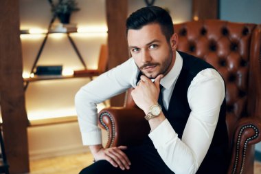 Yakışıklı genç adam modern lüks bir iç mekanda koltukta oturuyor ve poz veriyor. Moda portresi