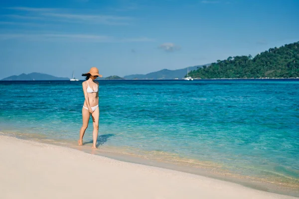 Frumoasă femeie tânără în bikini albi și pălărie de paie mergând pe plaja tropicală — Fotografie, imagine de stoc