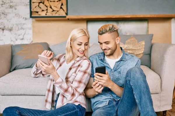 Sonriente pareja joven relajarse en el sofá con teléfono inteligente en casa —  Fotos de Stock