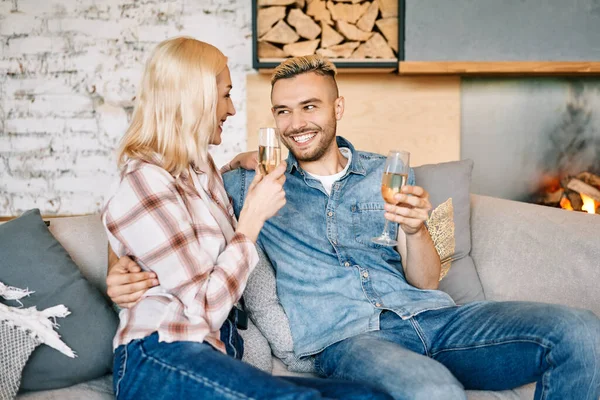 Joven pareja feliz bebiendo champán y sonriendo sentado en el sofá en su casa moderna —  Fotos de Stock