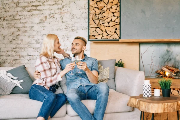Joven pareja feliz bebiendo champán y sonriendo sentado en el sofá en su casa moderna —  Fotos de Stock