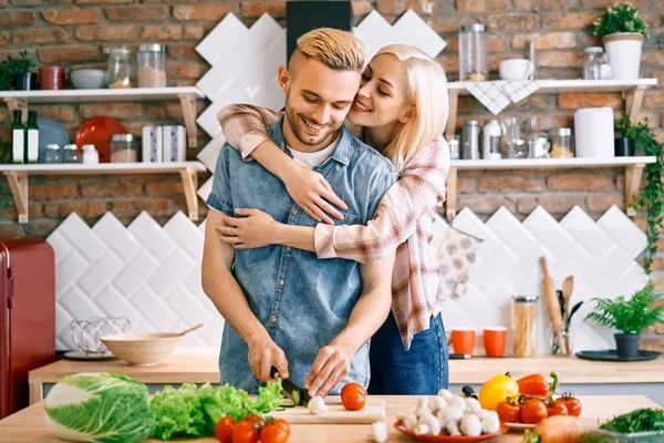 Улыбающаяся молодая пара готовит вместе вегетарианскую еду на кухне дома. Женщина обнимает мужчину — стоковое фото