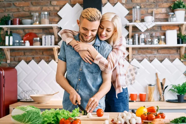 Leende unga par matlagning tillsammans vegetarisk måltid i köket hemma. Kvinnan omfamnar mannen — Stockfoto