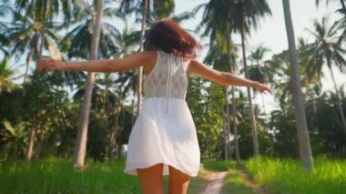 Beyaz elbiseli genç bir kadın palmiye ağaçları arasında kolları açık dönüyor.