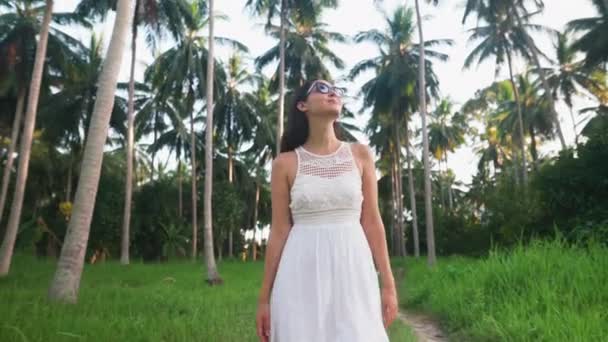 Jovem mulher de vestido branco caminhando na selva entre palmeiras na ilha tropical — Vídeo de Stock