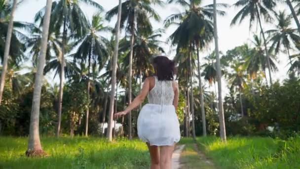 Счастливая молодая женщина в белом платье бегает в джунглях среди пальм на тропическом острове — стоковое видео