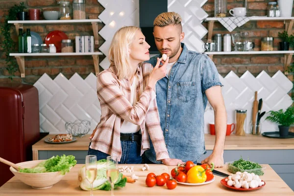 El joven se niega a comer verduras. Mujer alimentando al hombre con alimentos saludables mientras cocina en la cocina —  Fotos de Stock