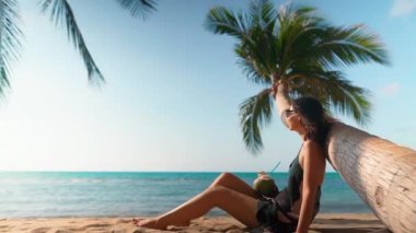 Güzel genç bir kadın tropikal sahilde, palmiye ağacının altında hindistan cevizi içeceğiyle rahatlıyor.