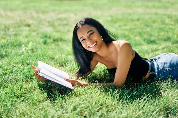Ładna afro amerykańska kobieta czyta książkę leżącą w trawie w parku — Zdjęcie stockowe
