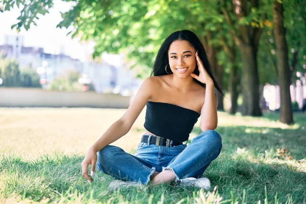 Pretty smiling afro american female posing in park sitting on green grass. — Stock Photo, Image