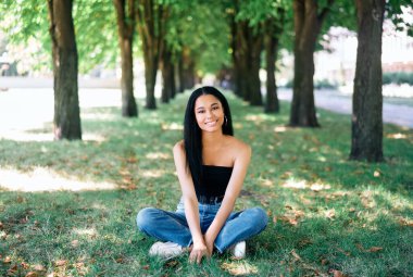 Parkta poz veren güzel, gülümseyen Afro-Amerikan kadın yeşil çimlerde oturuyor.