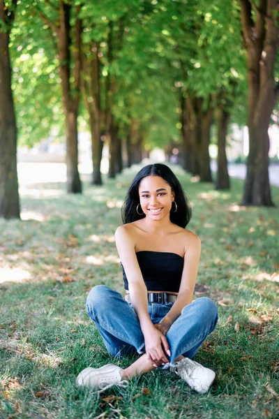 Muito sorridente afro-americano fêmea posando no parque sentado na grama verde — Fotografia de Stock