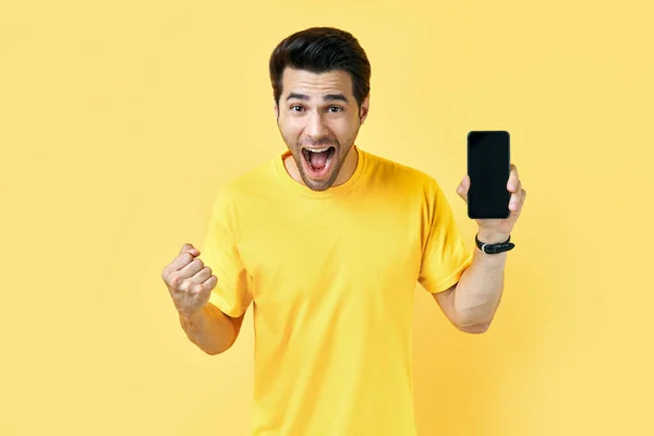 Emocionado hombre gritando sosteniendo el teléfono móvil y mirando a la cámara en el estudio amarillo —  Fotos de Stock