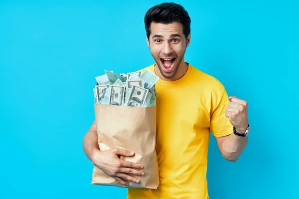 興奮した幸せな男ホールド紙袋フルのお金のスタックに隔離された青の背景. — ストック写真