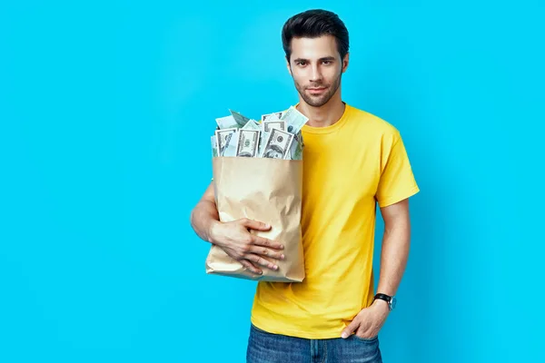 Handsome man hold paper bag full of stacks of money isolated on blue background — Stock Photo, Image