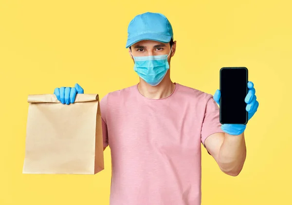 El repartidor sostiene el paquete de papel con comida y smartphone usando mascarilla facial y guantes como protección para COVID-19 —  Fotos de Stock