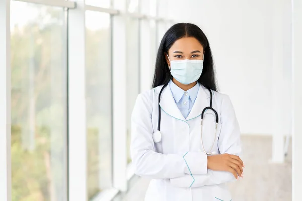Vrouwelijke arts draagt medisch masker met armen gekruist in het ziekenhuis achtergrond — Stockfoto