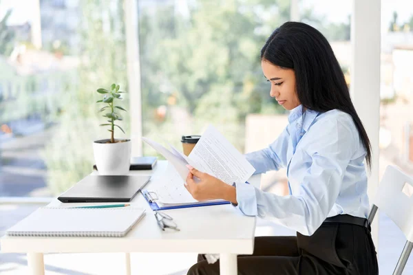 Hübsche schwarze Geschäftsfrau sitzt am Arbeitsplatz und liest Dokumente in ihrem modernen Büro — Stockfoto