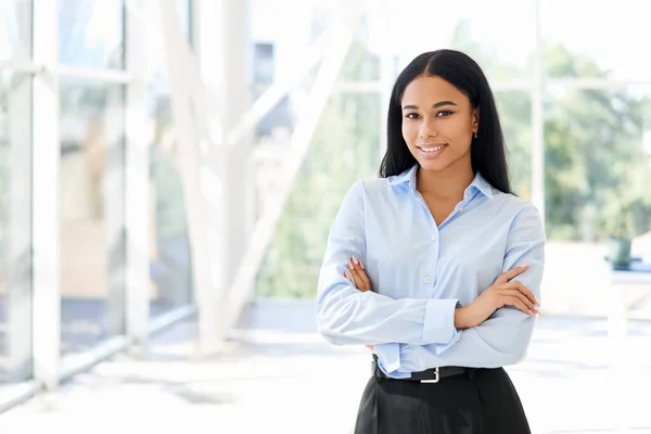 Улыбающаяся уверенная чернокожая деловая женщина, позирующая в современном офисе со скрещенными руками — стоковое фото