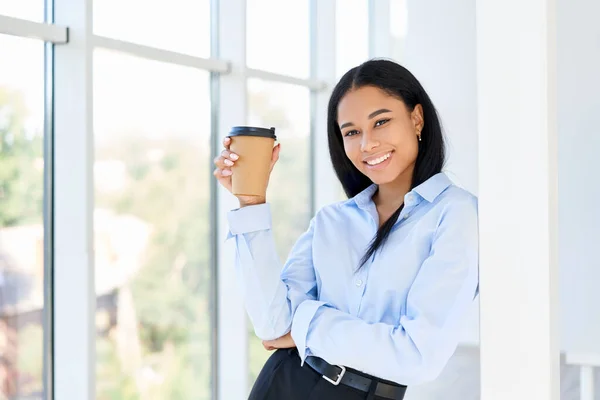 Šťastná černá obchodní žena s šálkem kávy v ruce během přestávky v moderní kanceláři — Stock fotografie