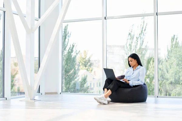 Красивая черная деловая женщина работает на ноутбуке сидя на кресле бобовый мешок с панорамным фоном окна — стоковое фото