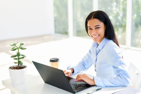 在笔记本电脑上工作的非洲裔美国女商人笑着坐在她的办公桌前 — 图库照片