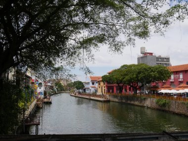 Malacca, Malezya--Şubat 2018: Melaka Nehri yaya köprüsünde görüldü, nehir kıyısında eski binalar ve büyük bir ağaç tarafından çerçeveli. 