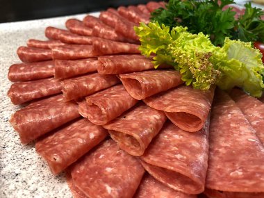 Taze yeşil sebzelerle süslenmiş tahtaya düzgünce yerleştirilmiş salam dilimleri.