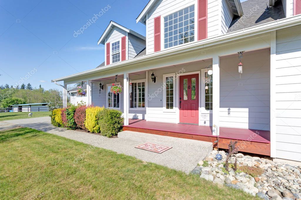 Nice home showcases a cozy covered porch with red front door and well kept lawn in the front yard on a bright summer day.