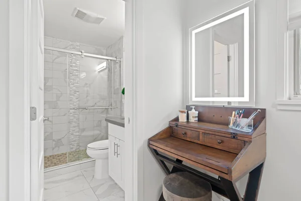 Closeup view of a makeup vanity with x base legs and a lighted mirror. Open door to an elegant master bathroom