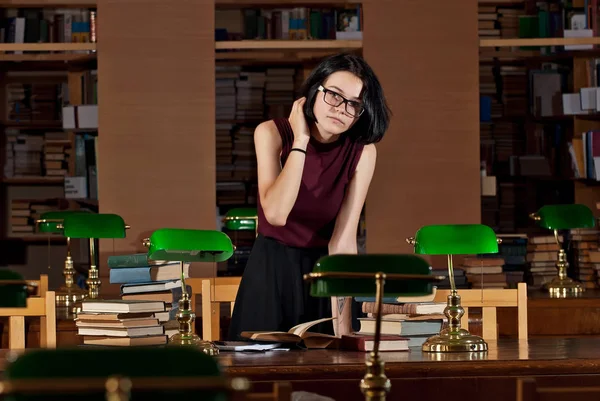 brunette in glasses at the table with books. Library with green lights on the tables. An old library with brown furniture.